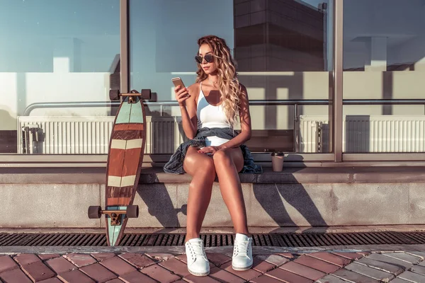 Beautiful girl athlete in casual clothes, summer city, skateboard, longboard, sits hand phone, reads application social networks online chat Internet, background of glass windows. Free space. — Stock Photo, Image