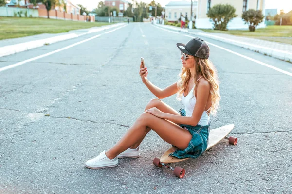 Indah bahagia gadis duduk skateboard, Ruang gratis untuk teks. Dia memegang smartphone tangannya, berbicara video call, aplikasi internet online. Mengambil gambar sendiri membuat selfie. Di kota musim panas . — Stok Foto