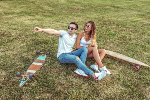 Glückliches, freundliches junges Paar, Frau und Mann, sitzen im Sommer auf Gras im Stadtpark. Skateboards, Longboard. Konzept-Teenager sind Teenager, trendy und stylisch in Freizeitkleidung. Erholung am Wochenende. — Stockfoto