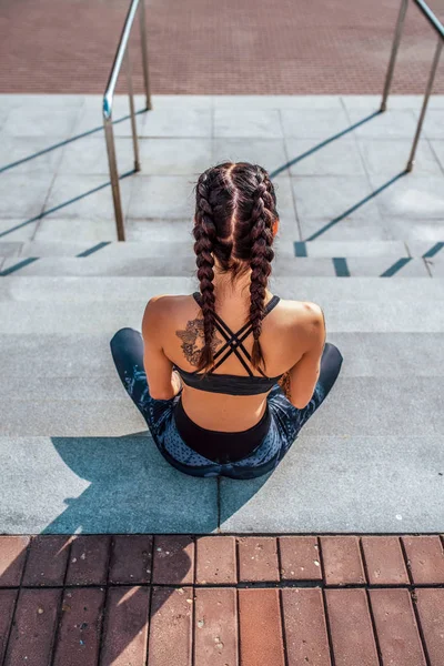 Atlet perempuan duduk di tangga di musim panas di kota. Santailah setelah latihan di udara segar, atasan legging pakaian. Kulit kecoklatan dengan tato, wanita modis dan bergaya. Tampilan belakang . — Stok Foto