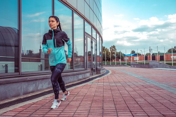 Athletic woman running morning run in summer city. Active lifestyle, girl motivation. Clothing leggings windbreaker. Free text space for motivation. Background of glass buildings and tiles in city.