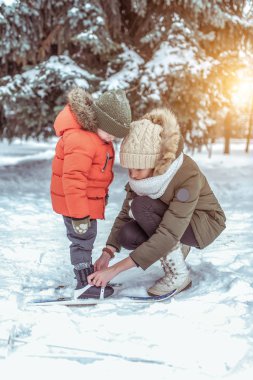 Genç bir anne çocuğunu 3-6 yaşında bir çocuk yardımcı olur, çocuk s kayaklar için giysiler giyiyor. Kayak ilk adımlar. Destek ve destek, sağlıklı yaşam öğrenme. Kışın şehir park ormanda.