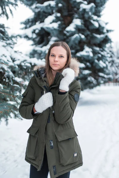 Uma menina fica no inverno no fundo de árvores cobertas de neve na floresta. Em uma jaqueta quente e luvas brancas. Aquece-se por um inverno frio o dia de fevereiro e janeiro . — Fotografia de Stock