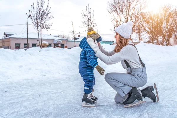 Mutter richtet ihren kleinen Hutsohn, 3-5 Jahre alt, Hut. im Winter in der Stadt auf einer öffentlichen Eisbahn. in warmer Kleidung gegen Schnee- und Schneeverwehungen und eine Eisbahn. Freiraum für Text. — Stockfoto