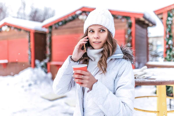 Poważną dziewczyną w białą kurtkę i ciepłą czapkę, rozmowy na telefon w zimie w mieście na świeżym powietrzu. W ręku trzyma szklankę napoju gorące śniadanie. — Zdjęcie stockowe