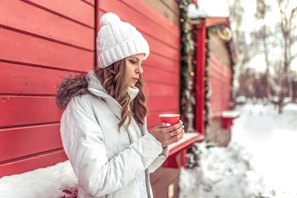 Krásná dívka se zahřívá o červený hrnek s horkou kávu čaj pít, zima venku. Odpočívající resort víkend, volné místo pro text. V bílém plášti a teplou čepici na pozadí červené oken. — Stock fotografie