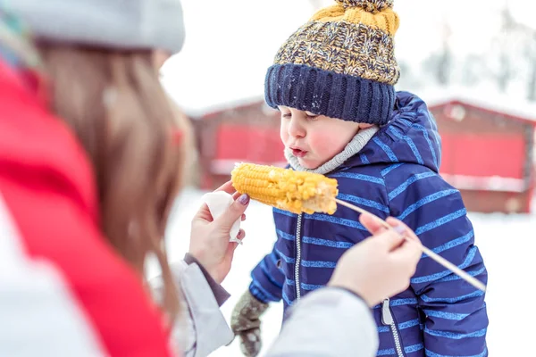 Сестра кормит младшего брата, сына 3-6 лет. В руках вареной кукурузы на палочке. Зимой в городе праздник, в выходные на отдыхе. Завтрак на свежем воздухе . — стоковое фото
