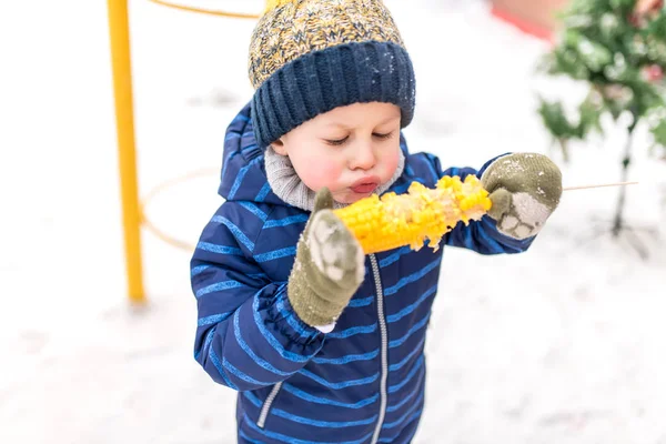 Маленький мальчик 4-5 лет, зимой в городе на свежем воздухе на фоне снега. Дуть на горячую вареную кукурузу. Обед и закуски по выходным в холодный праздничный день. В теплой одежде и шляпе . — стоковое фото