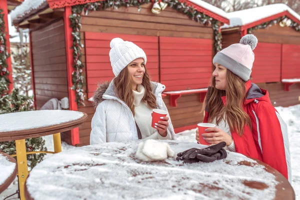 Dwie dziewczyny dziewczyny porozmawiać w zimie przy stole, holding kubki kawy i herbaty, gorący napój, śniadanie, obiad, przekąski, na świeżym powietrzu. Emocje radości zabawy weekend w ośrodku. — Zdjęcie stockowe