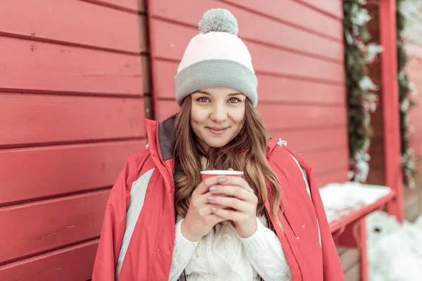 幸せな女が笑みを浮かべて、長い髪冬市の新鮮な空気を持つ少女。熱いカップの温めは、コーヒー紅茶を飲みます。赤いジャケットの温かみのある帽子。中庭の週末感情優しさ快適快適な時間を過ごす. — ストック写真