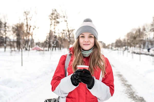 Seorang gadis di musim dingin di kota dengan latar belakang salju dan jalan, di tangannya memegang secangkir kopi, teh, hangat pada hari musim dingin. Emosi sukacita yang menyenangkan, beristirahat, memiliki camilan di alam . — Stok Foto