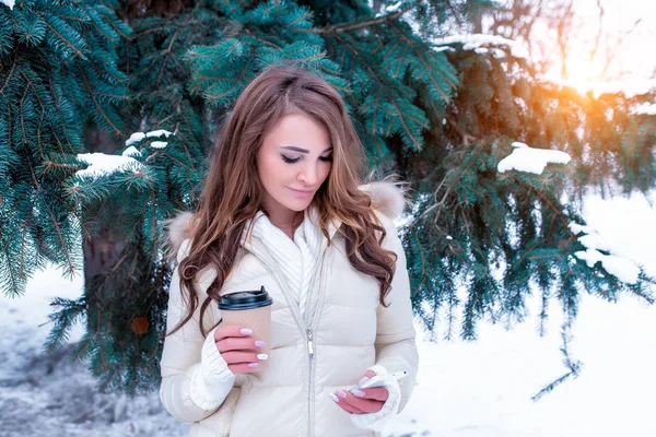 Hermosa mujer en invierno en el bosque. En su mano está un trabajador de la máquina con té de café caliente, un teléfono inteligente, lee y ve un chat, en línea en la aplicación de redes sociales . —  Fotos de Stock