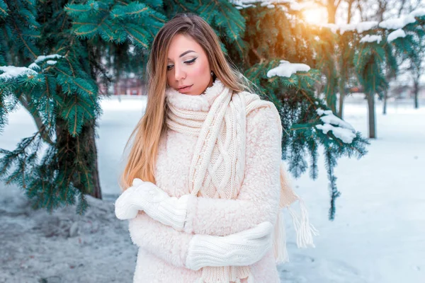 Ein schönes Mädchen mit langen Haaren, Winterwald vor der Kulisse von Schnee und grünen Tannen, wärmt sich in weißer warmer Daunenjacke und Fäustlingen. Gefühle des Komforts in der Natur, frische Luft genießen. — Stockfoto