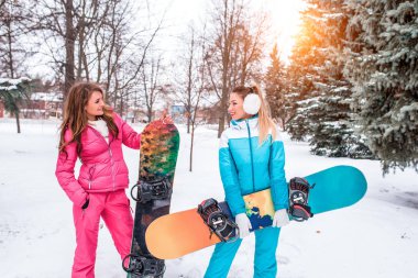 İki kız sporcular, bir kış ceket, dinlenme yamaç Kayak. Kar ağaçlar ve karla kaplı çam ağaçları fonunda. Mutlu snowboards elinde konuşuyor. Tatil hafta sonu.