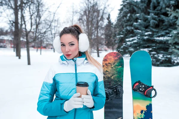 Ragazza in un cappello con cuffie, foresta invernale su sfondo di neve e alberi di abeti verdi. Nelle sue mani regge una tazza di caffè. Tuta blu, due snowboard, riposo al resort nel fine settimana . — Foto Stock