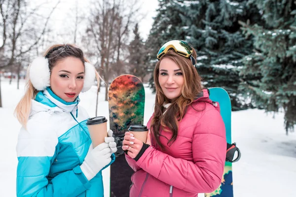 Dvě holky kamarádky stojí v zimě v lese na pozadí sněhu, zbytek resort na víkend na sjezdovce. Na pozadí snowboardy, v rukou šálků čaje kávy. — Stock fotografie