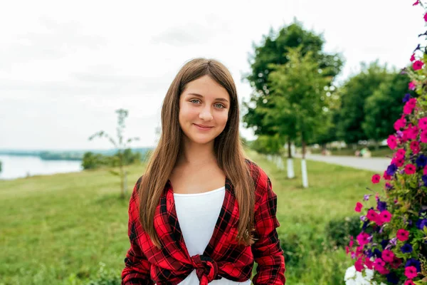 Ragazza adolescente 13-16 anni, felice sorridente si erge in estate nel parco su uno sfondo di fiori ed erba verde. Capelli lunghi grande sorriso. Camicia rossa, emozioni di gioia e divertimento . — Foto Stock