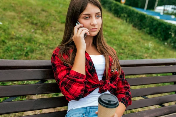 Adolescente 13-16 años de edad colegiala Chica sentada en el banco. En verano en la ciudad después de la escuela, los padres llaman, en sus manos una taza de té de café. Fin de semana de vacaciones en ciudad. Camiseta roja Camiseta y jeans . — Foto de Stock