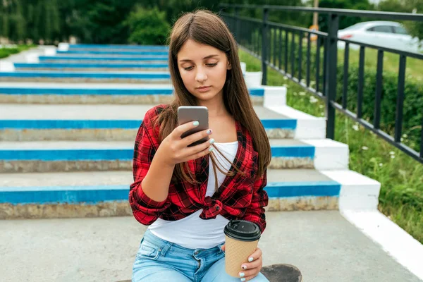 Studentessa adolescente 13-16 anni, seduta su uno skateboard. In estate in città dopo scuola, nello smartphone a mano, un bicchiere di bevanda. Legge e guarda l'applicazione internet, social network . — Foto Stock