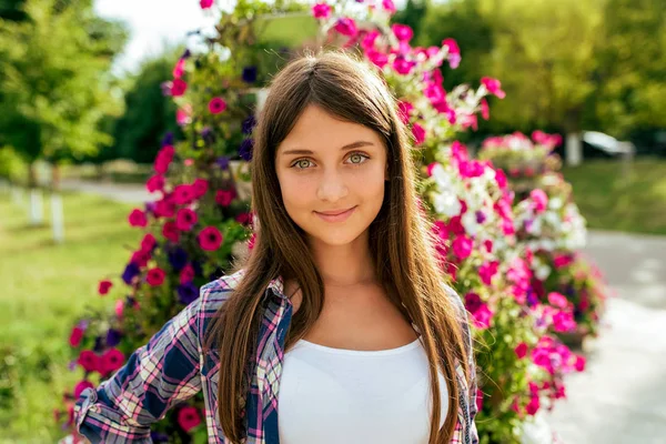 Schöne Mädchen Teenager 13-16 Jahre auf dem Hintergrund eines Blumenbeetes. glückliches Lächeln. im Sommer in der Stadt nach der Schule. Ferienwochenende in der Stadt. Hemd T-Shirt. Emotionen der Freude, des Spaßes, des Lebensstils. — Stockfoto