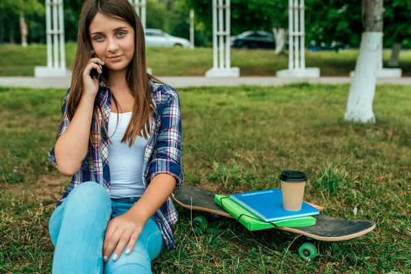 Девочка школьница, студентка 13-16 лет, сидящая в парке зеленая трава города. В руках телефон звонит родителям. Отдых за уровнем, возле скейтборда учебники ноутбуки стакан напитка, скейтборд. Пространство для текста . — стоковое фото