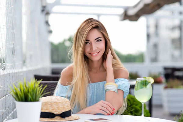 Šťastná usměvavá krasavice s dlouhými vlasy. Žena v letní kavárně na verandě restaurace. Sedí u stolu v modrých šatech, zblízka. Emoce radosti, radosti a pohodlí práce. — Stock fotografie