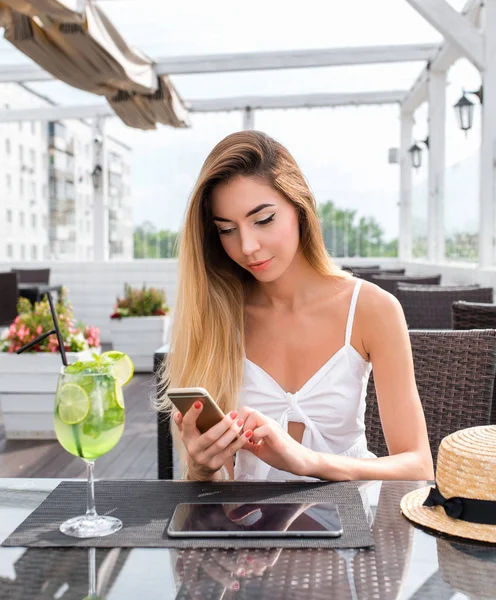 Schönes Mädchen in weißem Kleid, in einem Café sitzend, in der Hand ein Telefon, auf dem Tisch ein Glas mit Alkohol, ein Tablet. schreibt eine Nachricht online in Anwendung, Internet, sozialen Netzwerken Chat-Korrespondenz. — Stockfoto