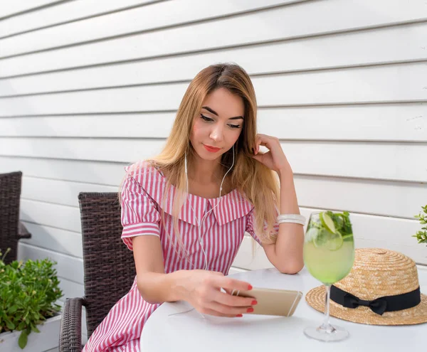 Beautiful woman watching video and listening to music headphones over the phone. The girl in summer cafe on veranda of restaurant. Sits at a table in his hand smartphone, music selection playlist.