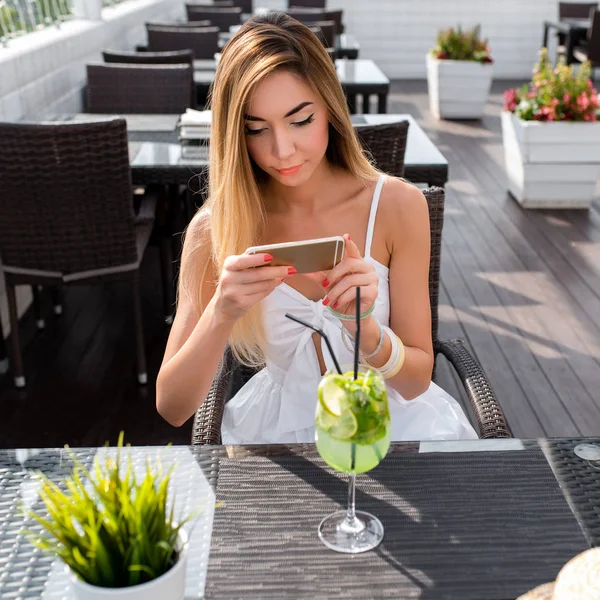 Girl photographs a glass of cocktail with a green lime drink. In the summer in cafe on veranda of the restaurant. In his hand a smartphone, an application to Internet, social networks, post, Like.