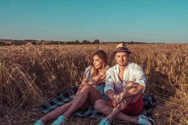 Ein junges Paar sitzt im Sommer in einem Weizenfeld auf einer Decke und entspannt sich an der frischen Luft. das Konzept romantischer Liebe, Segen und glücklicher Familienbeziehungen, Treffen und ein Date in der Natur. — Stockfoto