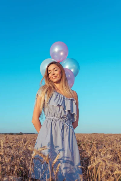 Menina feliz sorrindo, bela mulher fica no verão em um campo de trigo. Tem balões na mão. Descanse após o fim de semana, uma festa e aniversário na natureza. Cabelo longo com pele bronzeada . — Fotografia de Stock