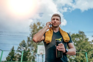 Erkek bir atlet, egzersiz, aramalar telefon, elinde tutuyor sonra bir shaker, bir şişe su, boyun etrafında bir havlu yaz şehir. Sert bir çalışmadan sonra dinlenme. Aktif yaşam tarzı, egzersiz doğada.