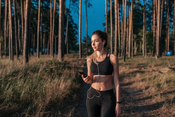 Beautiful tanned girl walk in summer park, background of trees. Sportswear leggings top. In hand phone, listening music motivation, sports before training. Online playlist application on smartphone.