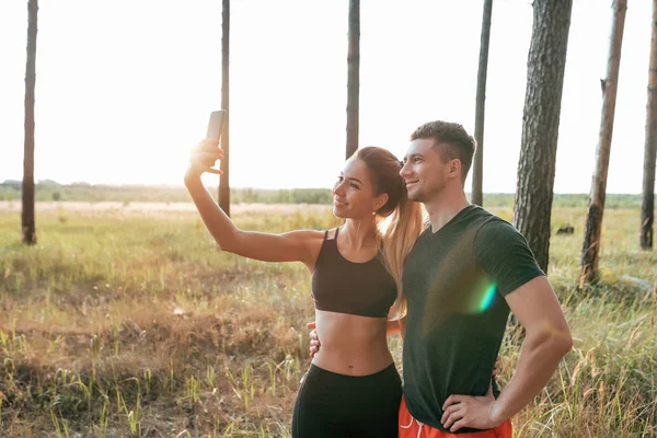 Młoda para, mężczyzna i Kobieta, lato park, są fotografowane przez telefon, szczęśliwy, uśmiechnięty, przed jogging, sportów na świeżym powietrzu, aktywny tryb życia. Sportowe emocje radości i zabawy. — Zdjęcie stockowe