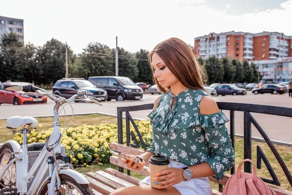 Una chica se sienta en un banco en verano en la ciudad, sosteniendo un teléfono en la mano con una taza de té de café. Bolsa de bicicleta. Lee un mensaje en la aplicación en línea, redes sociales en línea . — Foto de Stock