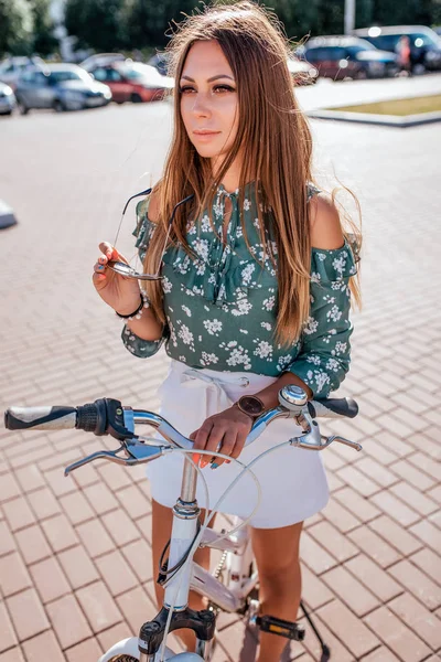 Загорелая девушка с длинными волосами стоит летом в городе на велосипеде, в руках солнцезащитных очков. Вид вдаль, летняя прогулка по городу на велосипеде . — стоковое фото
