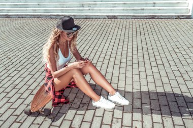 Kız bir kaykay üzerinde oturur, longboard, onun elinde bir smartphone, yazar ve bir iletiyi Internet üzerinde online sosyal ağları uygulama okur. Yaz aylarında şehrin kaldırım kutunun üzerindeki.