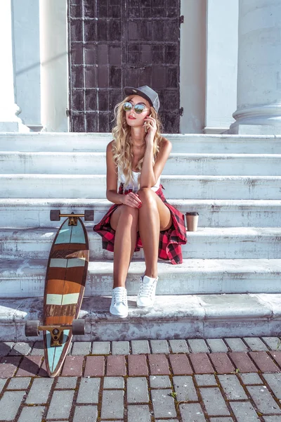 Ein schönes Mädchen sitzt auf den Stufen, im Sommer in der Stadt, telefoniert, neben einem Skateboard, einem Longboard, einer Tasse Kaffee und Tee. vor dem Hintergrund von Treppen und weißen Säulen. — Stockfoto