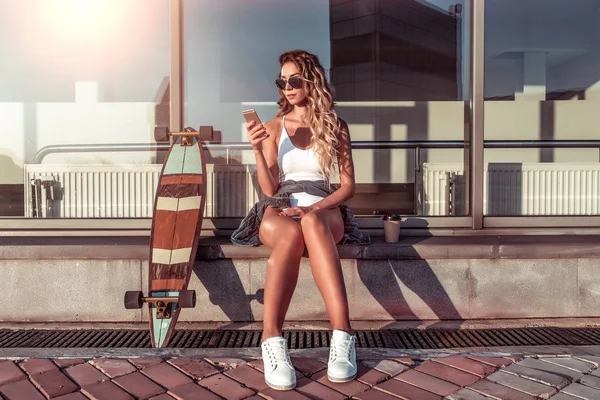 Chica se sienta edificio de fondo de la ciudad de verano, su teléfono de mano aplicación en línea Internet, monopatín, longboard. Espacio libre. El concepto de moda, estilo, ropa moderna para los jóvenes relajarse . — Foto de Stock