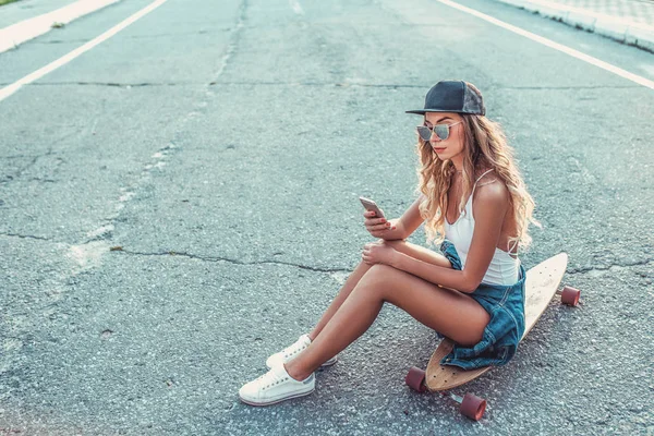 Dívka s dlouhými vlasy sedí v létě ve městě na skateboard, longboard. Prázdninový víkend, mladý módní a stylové. Koncepce životního stylu, módní ženy. Volné místo pro text. — Stock fotografie