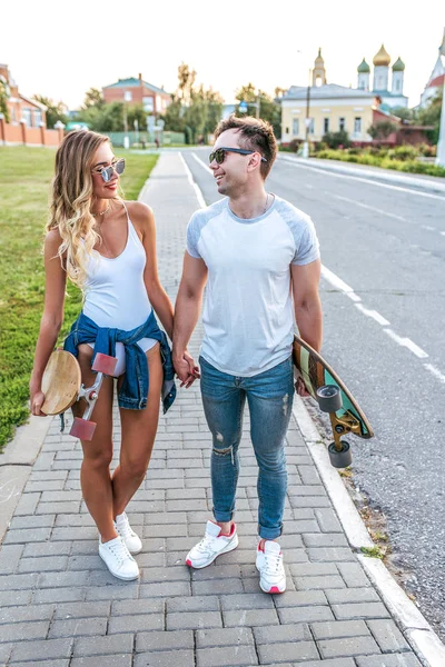 Pareja joven hombre y mujer caminan en el verano en la ciudad, en las manos de patinetas, longboard. Feliz sonrisa jugando divertido. Caminar en el fin de semana, relaciones amorosas familia fuerte . — Foto de Stock