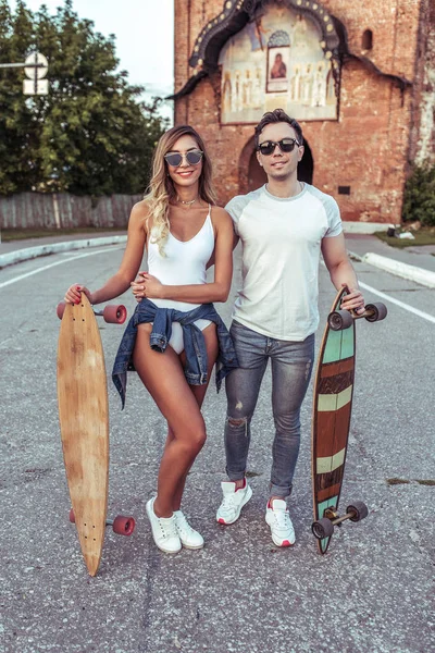 Krásná mladá dvojice muž a žena chůze letní město, ruce skateboard, longboard. Spokojený úsměv pózuje, baví. Chodit na víkend, rádi silné rodinné vztahy. Emoce, pozitivní a milující. — Stock fotografie