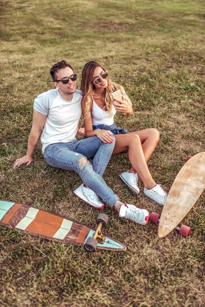 Adam kadın ilişki yaz şehir çekici çimen, oturup telefon selfies alarak, Resimler. Longboard kaykay, güneş gözlüğü. Duygular sevinç eğlenceli, temiz hava hafta sonları rahatlatıcı. Online görüntülü arama uygulaması.