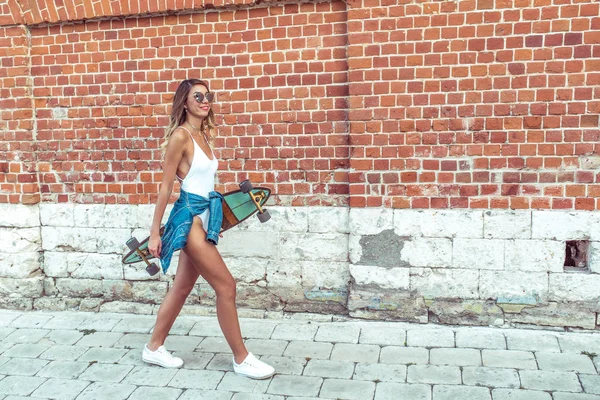 Chica en traje de cuerpo blanco con jeans, camina en verano en la ciudad en el fondo de una pared de ladrillo. Feliz sonrisa, piel bronceada. Espacio libre para texto . — Foto de Stock