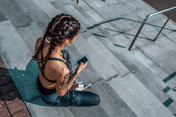 Κορίτσι που κάθεται το καλοκαίρι στην πόλη για τα βήματα, ανάπαυση μετά από μια προπόνηση. Στο χέρι του ένα smartphone με τα ακουστικά, βλέποντας μια εφαρμογή στο Internet, σε απευθείας σύνδεση εκπαίδευση σε κοινωνικά δίκτυα. Ελεύθερου χώρου. — Φωτογραφία Αρχείου