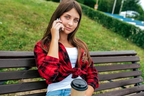 Een meisje zit op een bankje en maakt telefoongesprekken, een school meisje roept ouders op een smartphone, in de zomer in de stad in haar hand een kopje koffie in een rood hemd. — Stockfoto
