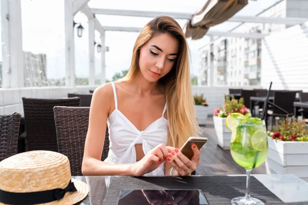 Ein Mädchen in einem Café im Sommer in der Stadt, schreibt eine Nachricht am Telefon, Online-Anwendung im Internet. Mittagspause im Restaurant auf der Veranda. lange Haare weißes Kleid. — Stockfoto