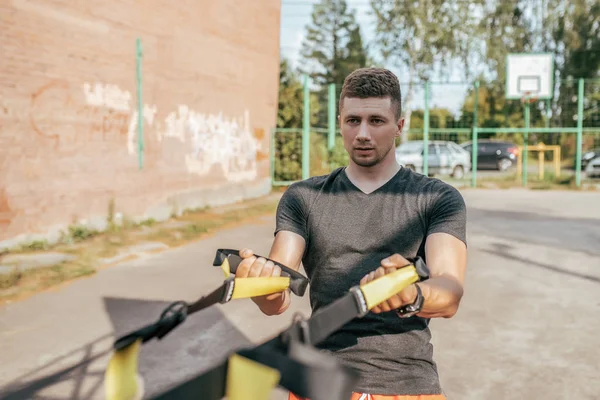 Männliche Sportler, im Sommer in der Stadt auf dem Sportplatz. Fitness mit Gummischlaufen, Gurten für Muskeln. aktiver Lebensstil, Bizepstraining. Fitness in der Natur. Freizeitkleidung. — Stockfoto