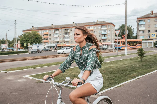 Vacker flicka i rörelse rider en cykel i staden. På sommaren på frisk luft. Livsstil i staden. Känslor av komfort och säkerhet. Grön blus och vit kjol shorts .. — Stockfoto