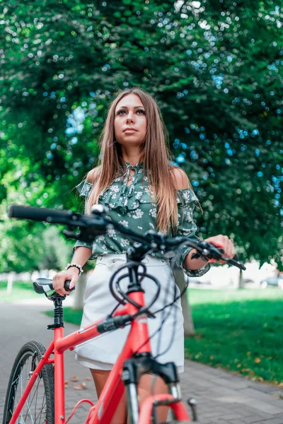 O fată frumoasă stă lângă o bicicletă roșie pe fundalul copacilor din parc. Vara pe aer curat. Stil de viață în oraș. Emoții de confort și siguranță. Bluza verde si fusta scurta alba . — Fotografie, imagine de stoc